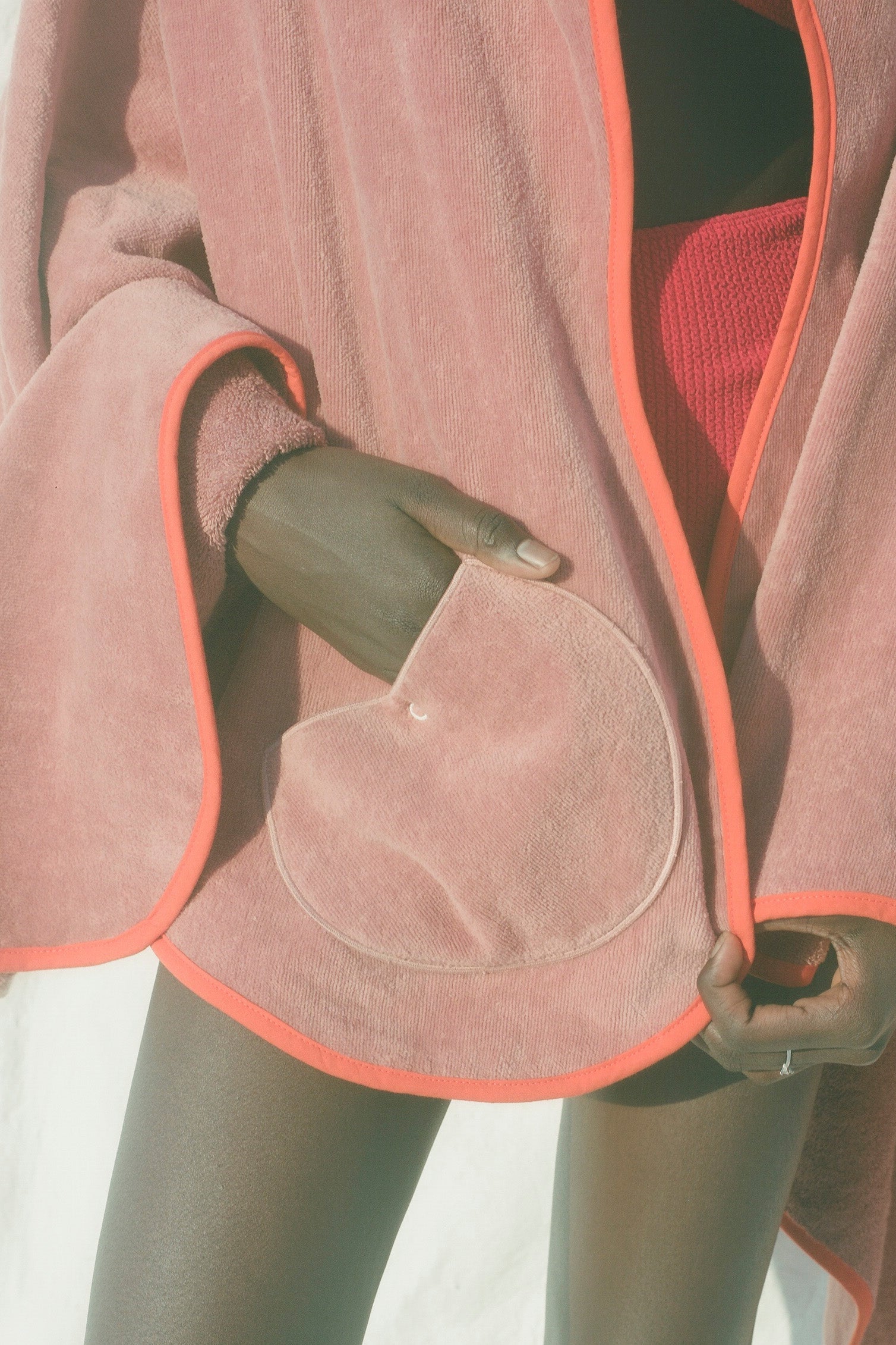 A close-up of a dusty pink plush bath towel draped over a model, featuring a bright orange-pink piping border and a large heart-shaped pocket.