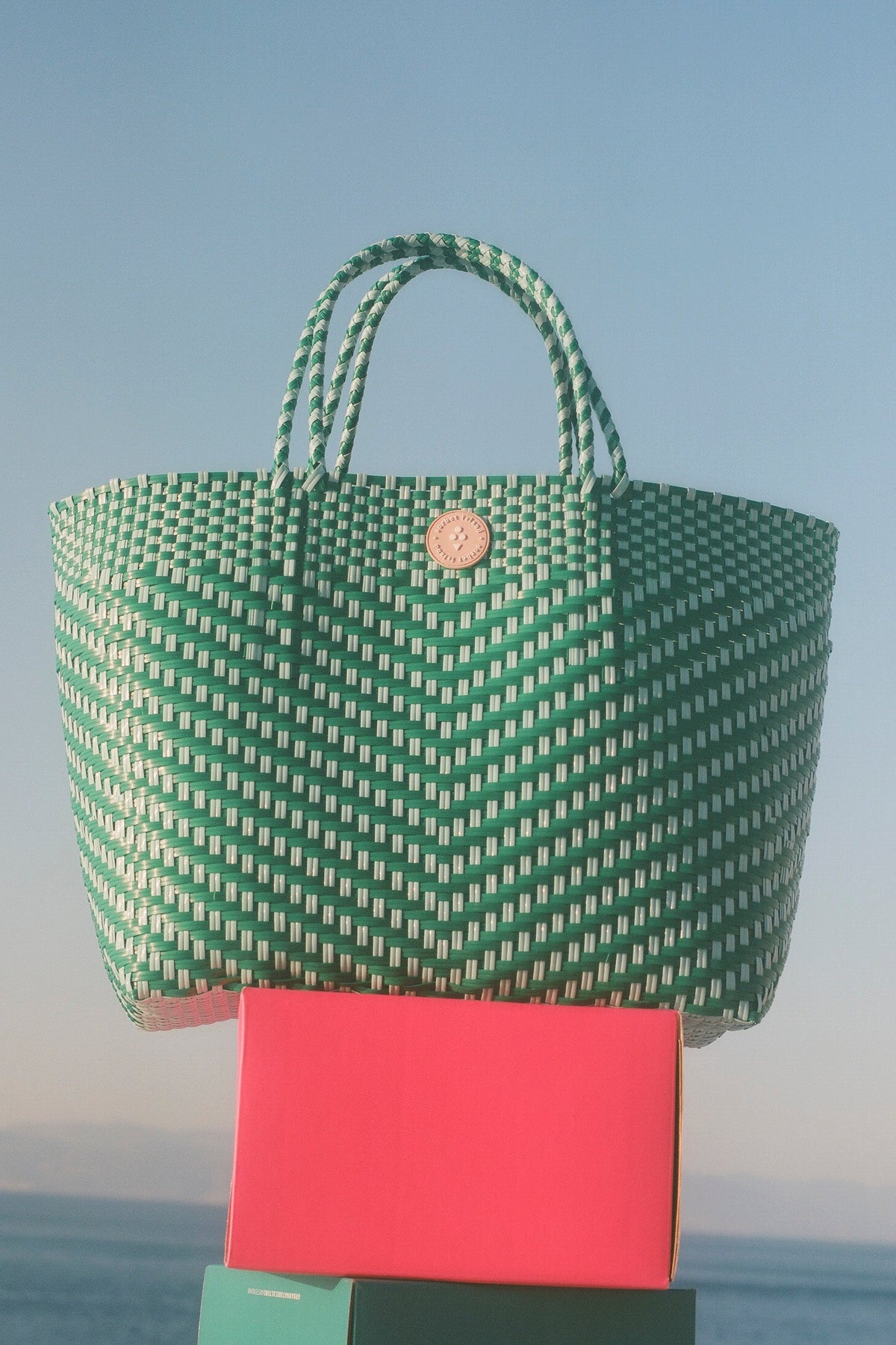 A green and white woven tote bag with rope handles and a round gold-tone logo button, displayed outdoors on top of a bright pink box with a blue sky and sea in the background.
