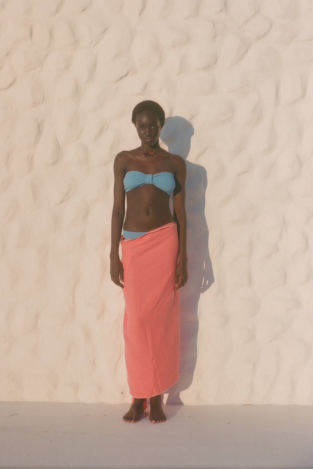 A model lies on a white sandy beach wearing a light blue bikini top and a coral-pink sarong wrap.