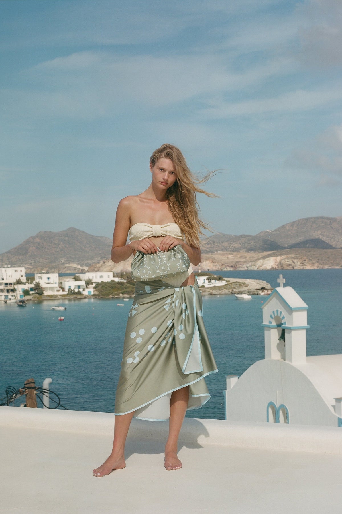A woman stands barefoot on a white wall overlooking a blue sea and mountains, wearing a strapless sage green wrap dress with white floral patterns.