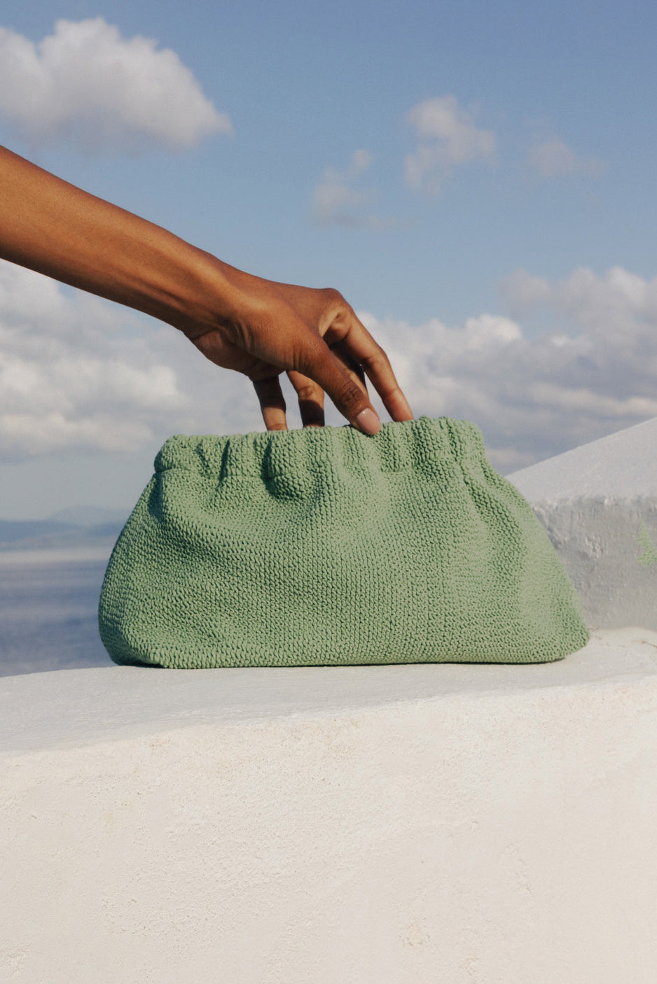 A sage green textured fabric clutch bag with a gathered top handle, held by a person's hand against a scenic background of a white wall and blue sky.