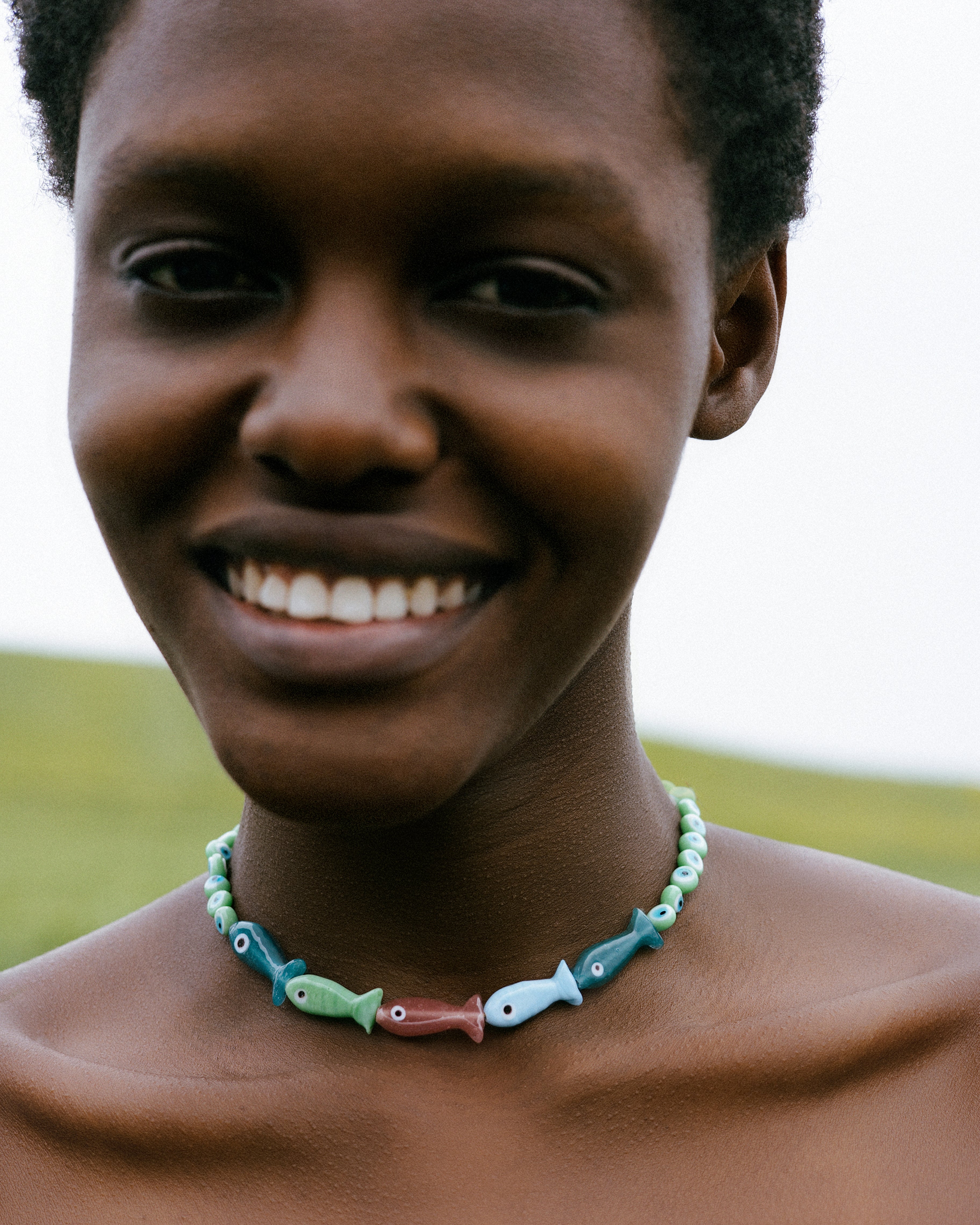A smiling person wearing a beaded necklace with fish-shaped beads in light blue, green, and red, photographed outdoors against a soft blurred background.