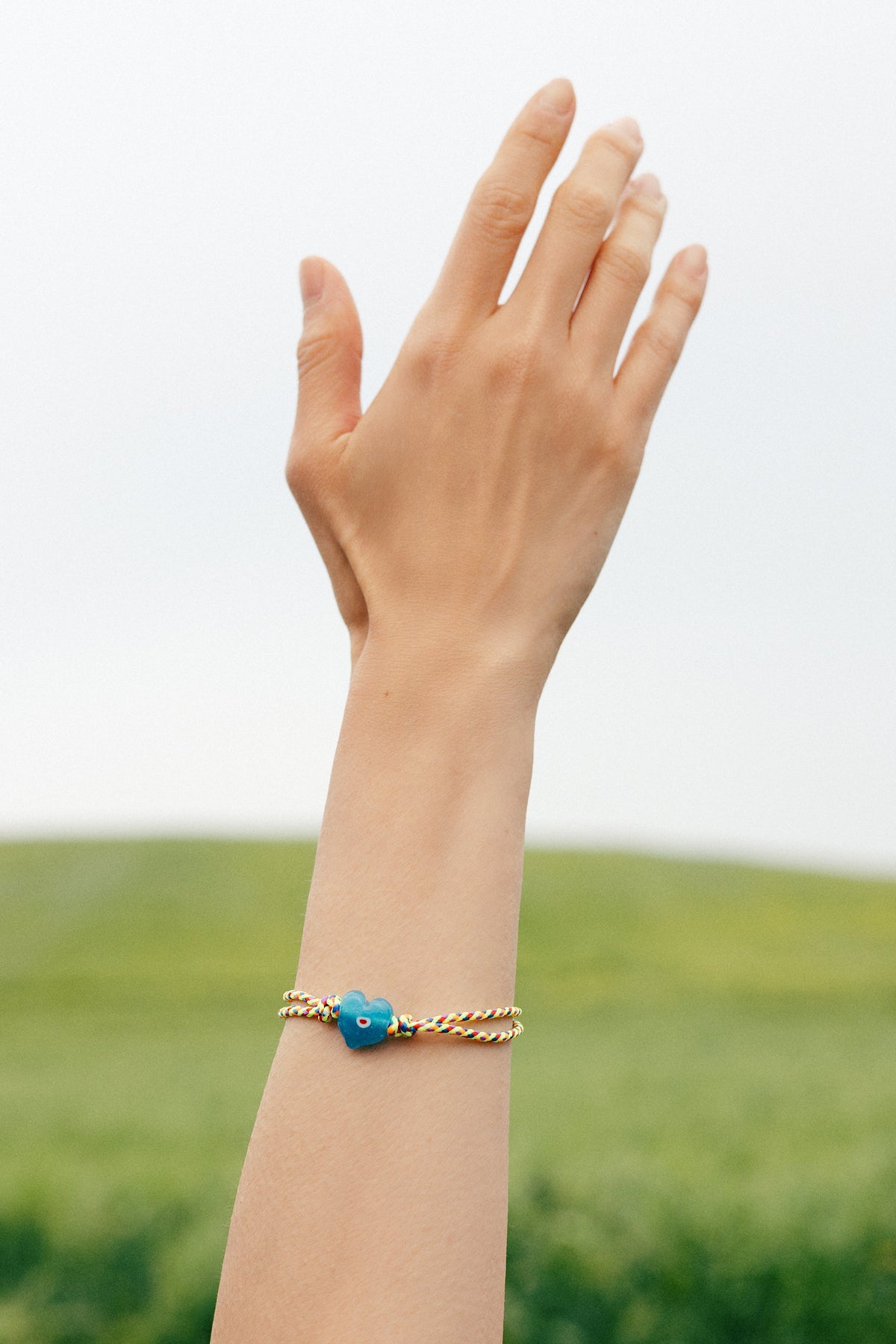 A gold chain bracelet with a small blue flower charm worn on a person's wrist, shown against a soft-focus outdoor background of green grass and a bright sky.