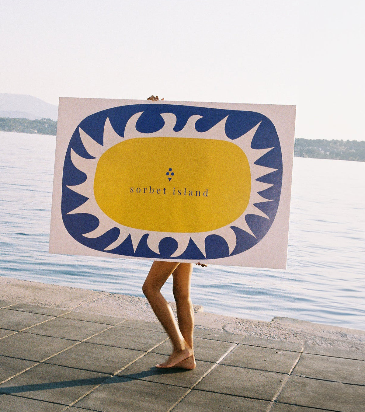 Person holding a large sign with a sun design and 'Surfet Island' text by a body of water.