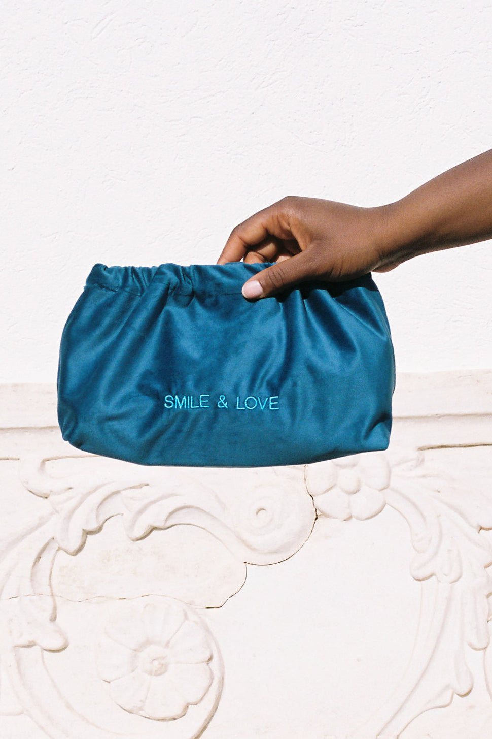 A hand holding a teal blue satin drawstring pouch with the text 'SMILE & LOVE' printed in light blue on the front, set against a white textured wall and a white carved stone ledge.