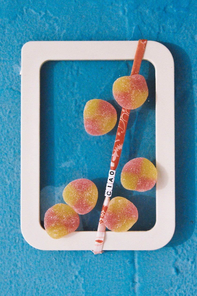 A white rectangular frame containing several pink and yellow gummy candies and a red-and-white striped paper straw with the word 'CIAO' printed on it, set against a bright blue textured background.
