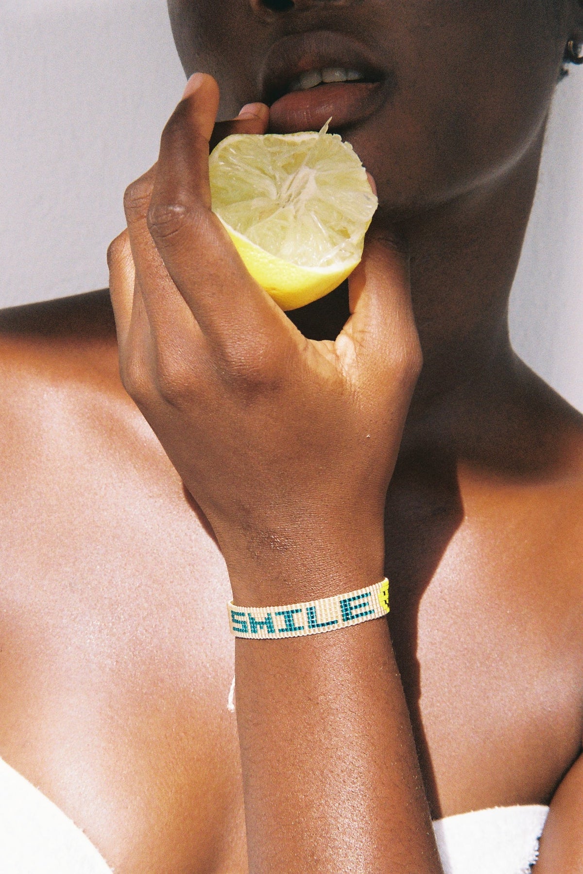 A close-up lifestyle shot of a woman wearing a beaded bracelet that spells 'SHILE' in yellow beads, holding a lemon half near her mouth.