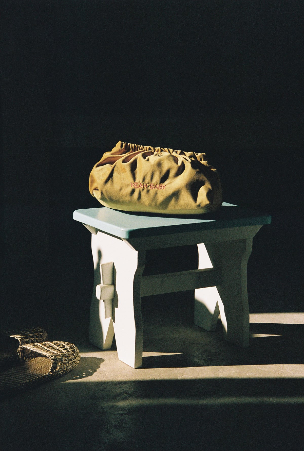 A tan canvas drawstring pouch bag with pink embroidered text sits on a white wooden stool against a dark background.