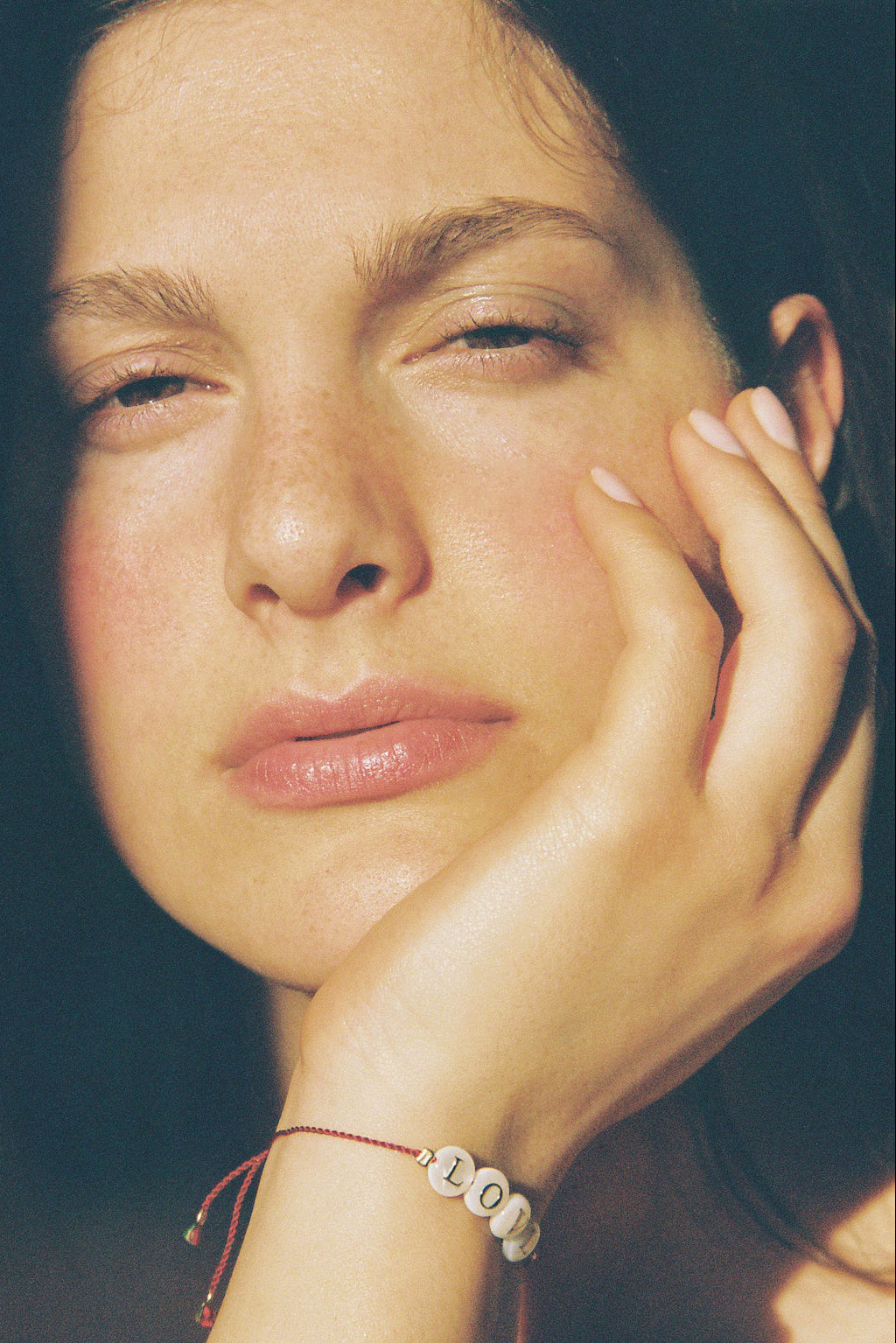 A close-up portrait of a woman resting her head on her hand, wearing a thin red cord bracelet with white beads spelling '100' on her wrist.
