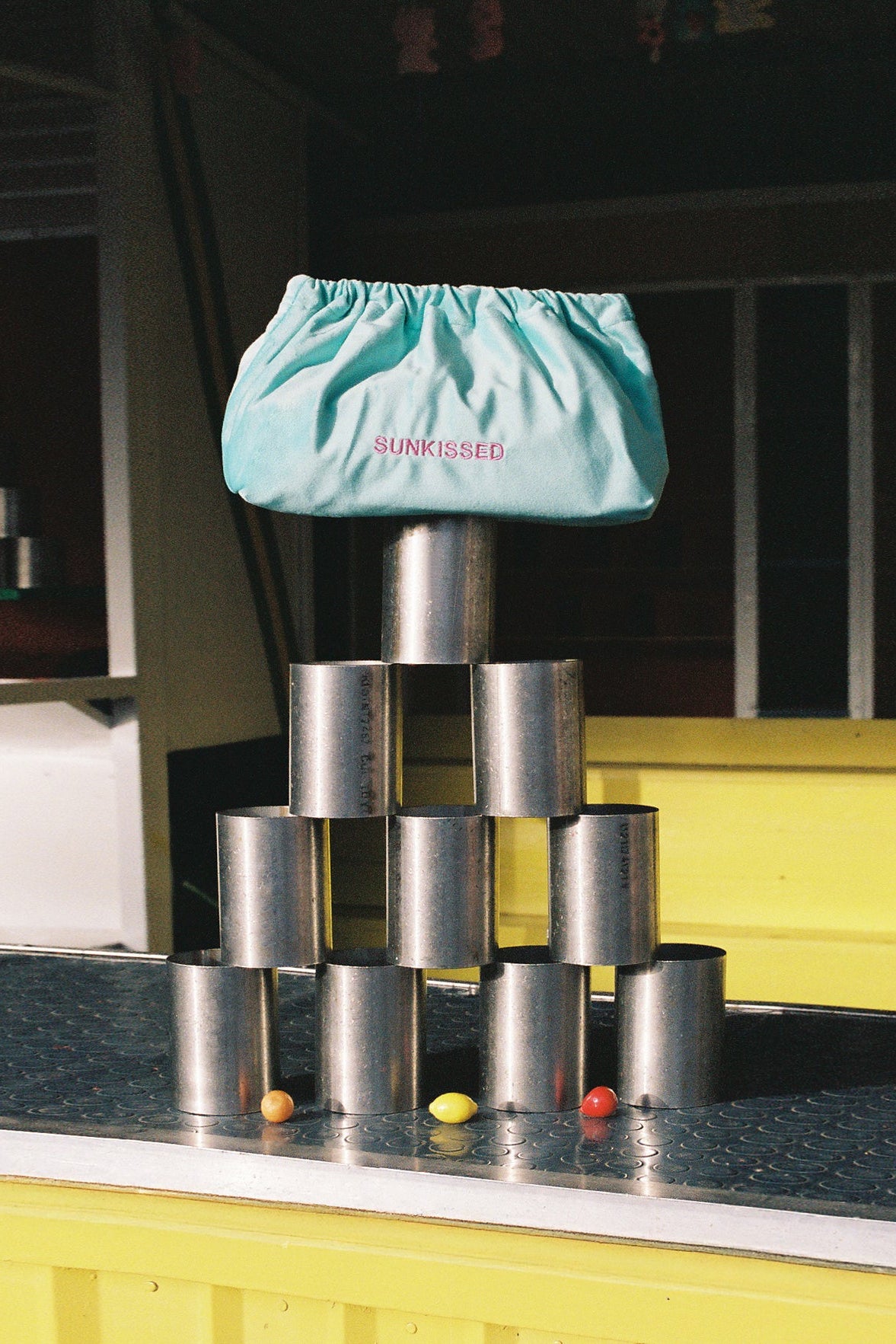 A light blue drawstring pouch with the word 'SUNKISSED' printed in pink on the front, balanced on top of a stack of metallic silver cans on a counter.