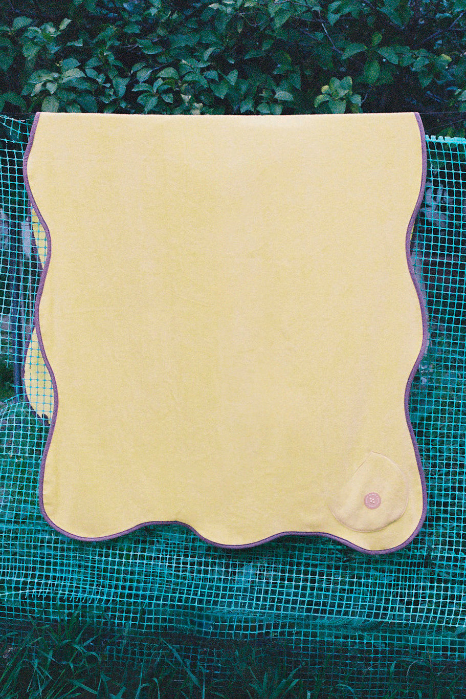 A pale yellow rectangular mat with wavy scalloped edges and a small circular pink detail in the lower right corner, displayed outdoors on a green mesh screen with foliage in the background.