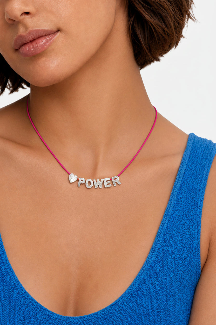 A close-up of a model wearing a silver 'POWER' necklace with a small heart on an orange cord, paired with a bright blue top.