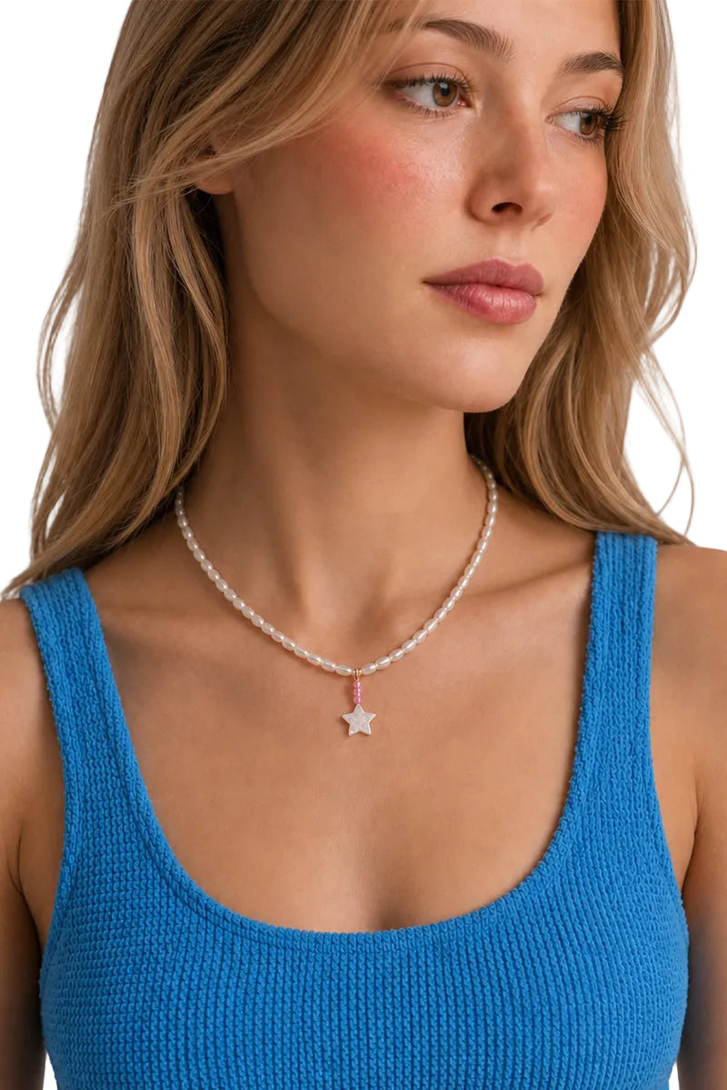 A close-up of a woman wearing a delicate white pearl necklace with a small silver star pendant, paired with a bright blue knit tank top.