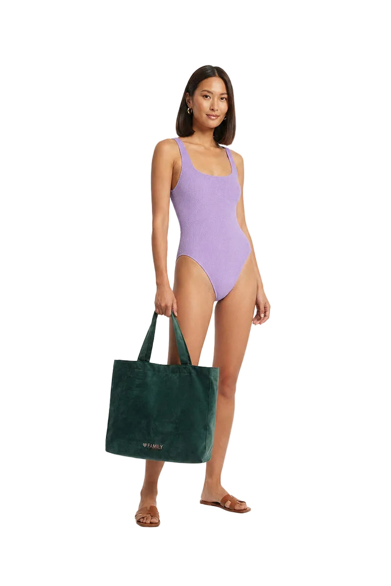 A model wearing a lavender one-piece swimsuit holds a dark green velvet tote bag with 'WANDERERS' printed in gold on the front, set against a plain light gray background.