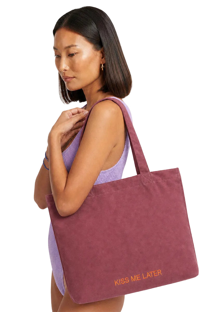 A woman wearing a lavender swimsuit holds a dusty rose canvas tote bag with the words 'KISS ME LATER' printed in orange on the bottom right. The background is a clean, light off-white studio backdrop.
