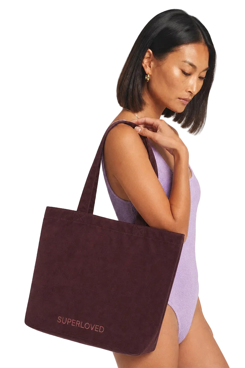 A woman wearing a light purple swimsuit carries a dark burgundy canvas tote bag with 'SUPERLOVED' printed in pink on the front, set against a plain light gray background.