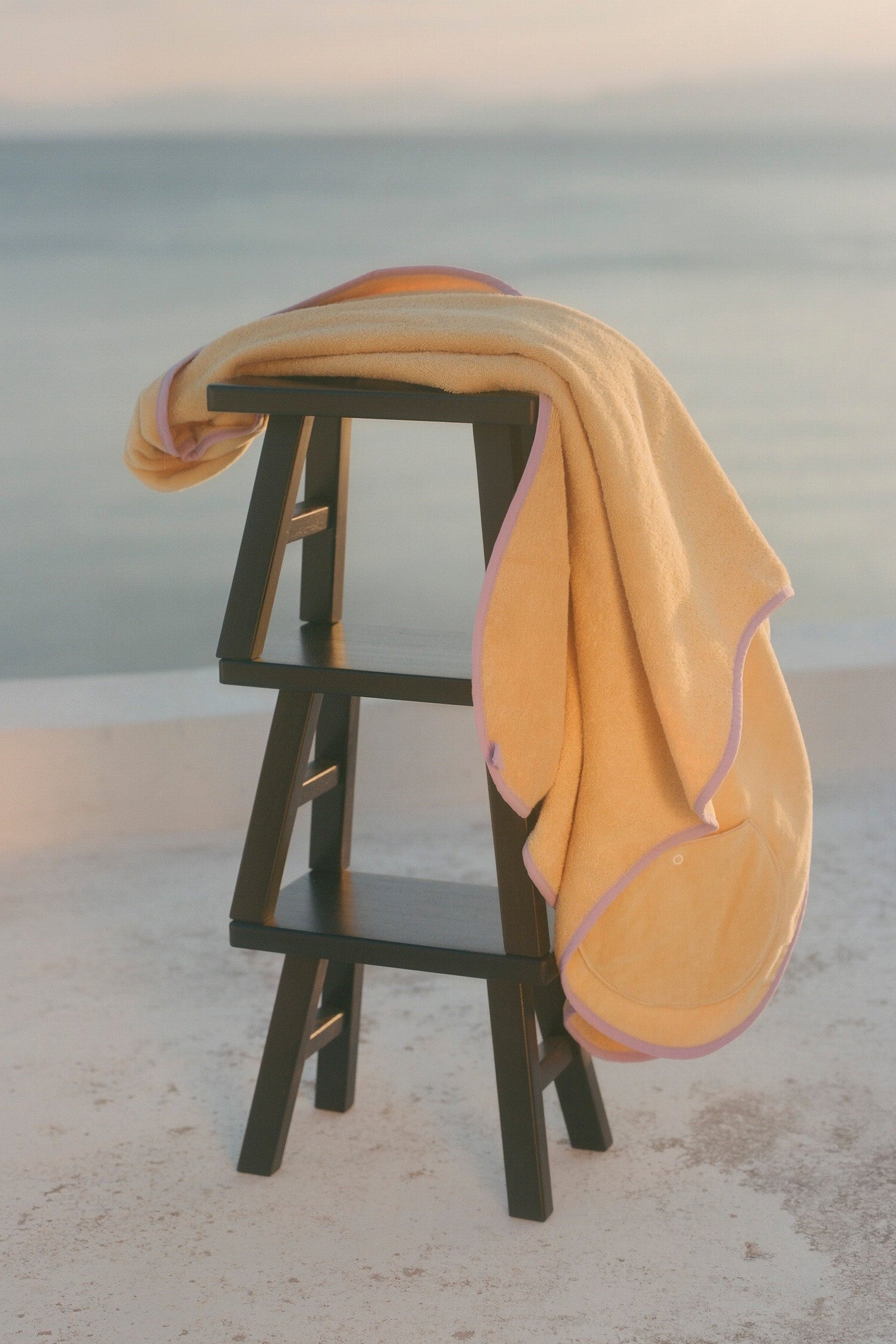 A soft pastel yellow towel with a lavender-purple border draped over a dark wooden stool, set against a blurred beach or seaside background at sunset.