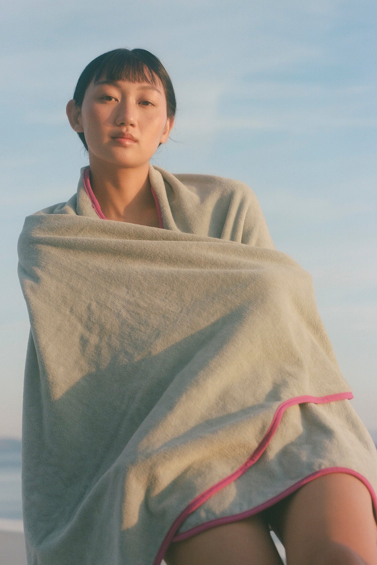A woman wrapped in a light gray beach towel with pink trim, sitting outdoors against a soft blue sky and distant water background.
