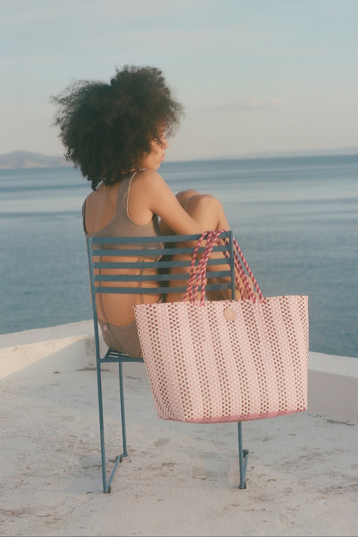 A woman sits on a blue metal chair by the sea, viewed from behind, with a large pink woven tote bag resting on the chair. The background shows a calm blue ocean and sky.