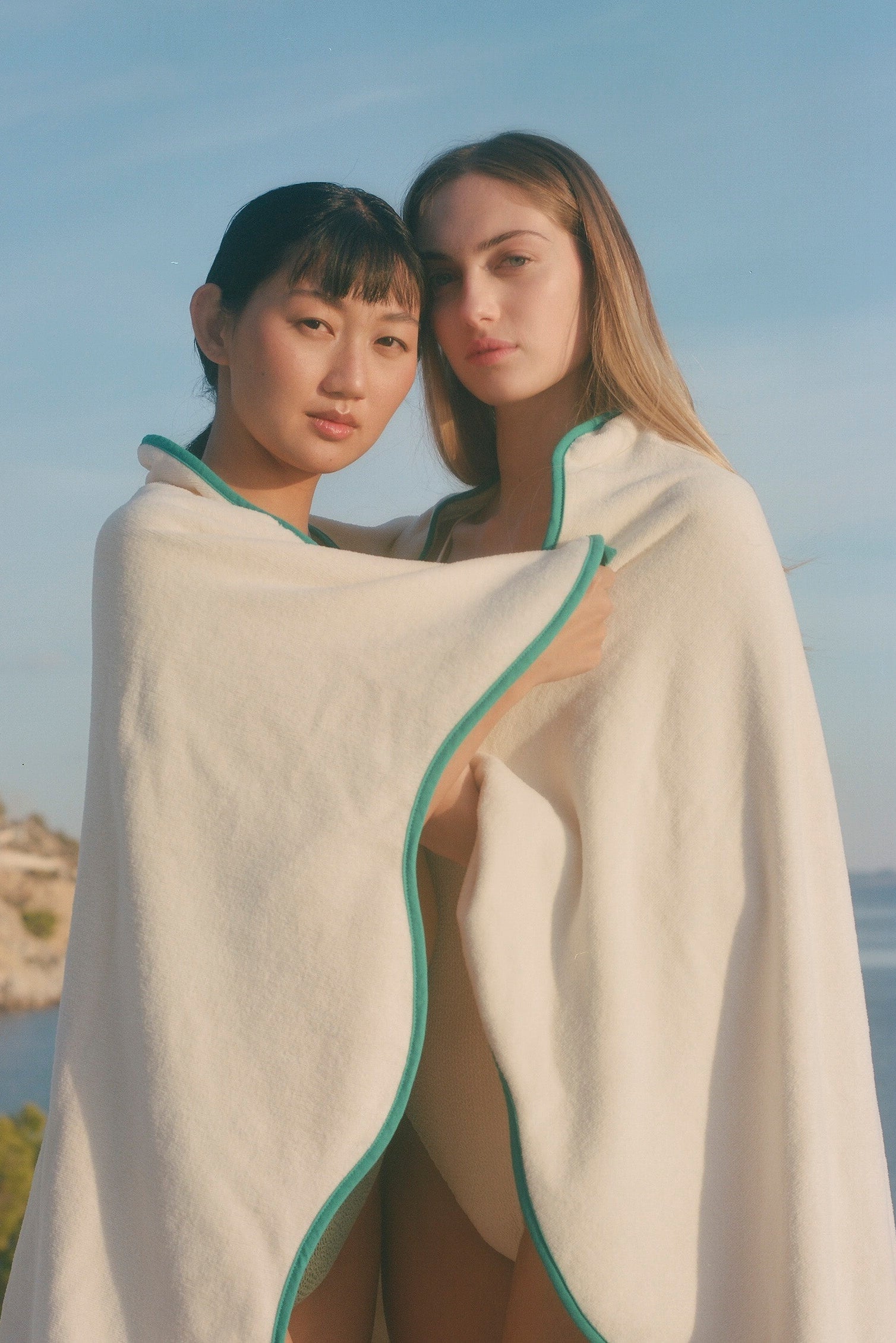 Two women standing outdoors by the sea under a blue sky, wrapped in a large off-white towel with a teal-green border.