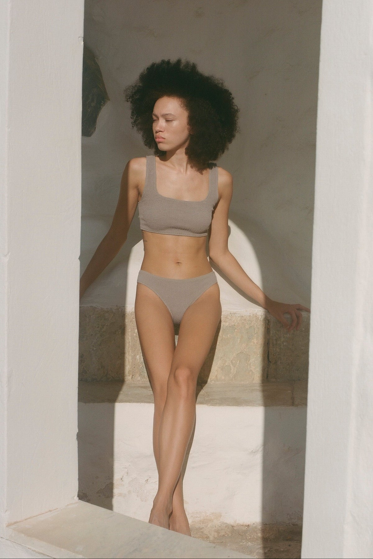 A woman with curly dark hair models a matching beige two-piece swimsuit consisting of a scoop-neck bikini top and high-cut bikini bottoms, standing in a sunlit white stone alcove.