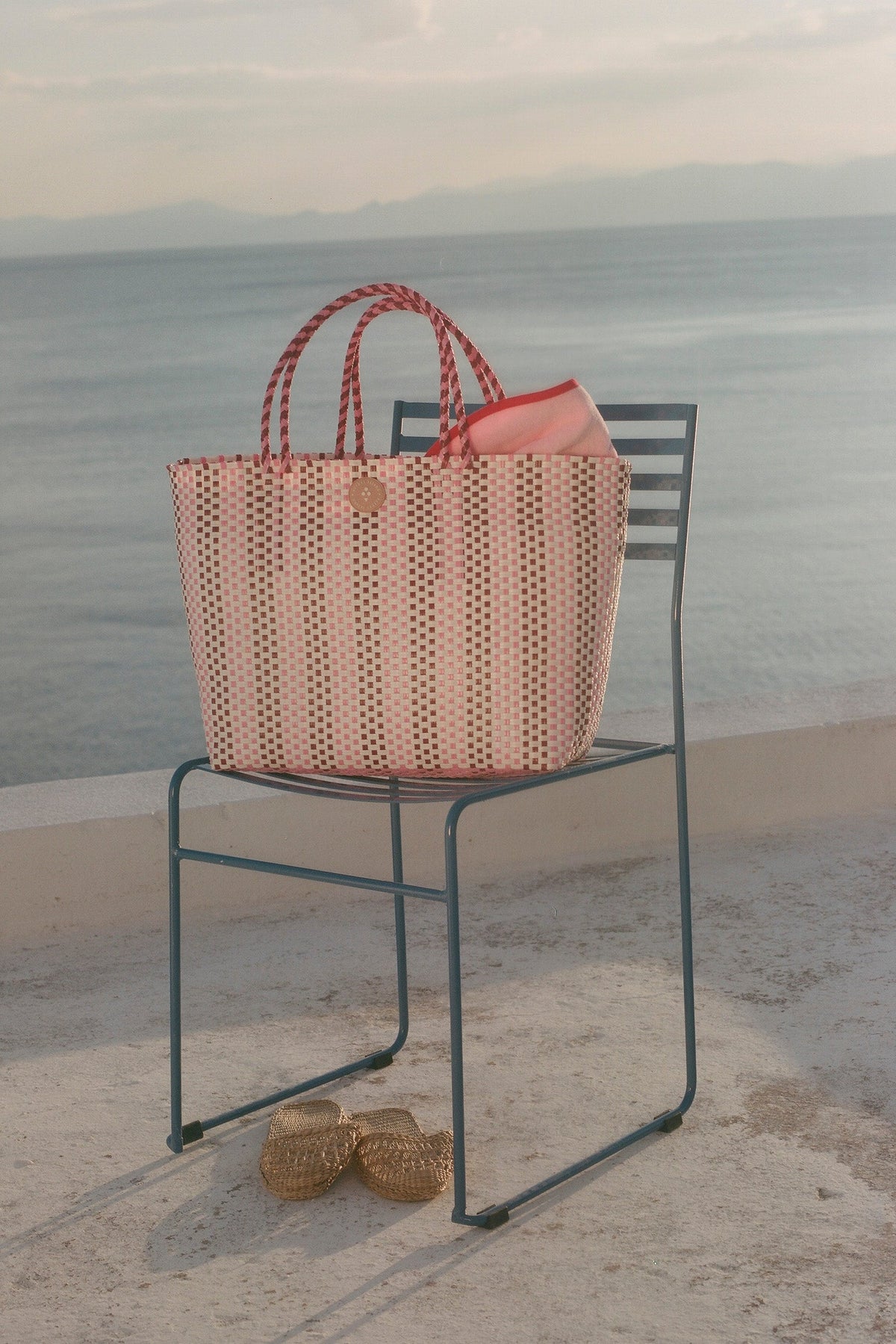 A woven straw tote bag with red-and-white braided handles sits on a metal chair by the sea. A coral pink pouch is tucked inside, and a pair of woven sandals rests on the ground below.