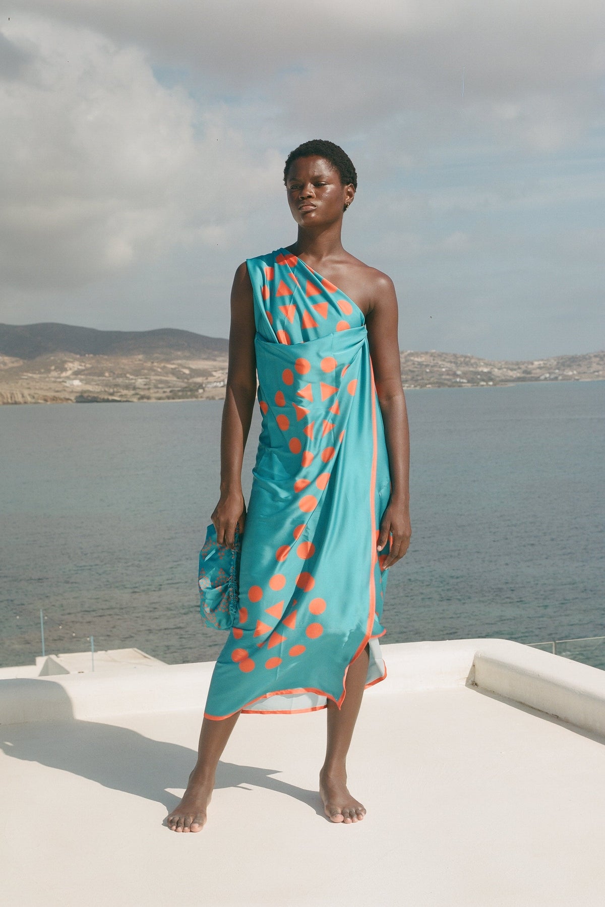 A model stands on a white deck overlooking a sea and distant hills, wearing a one-shoulder turquoise dress with orange polka dots.
