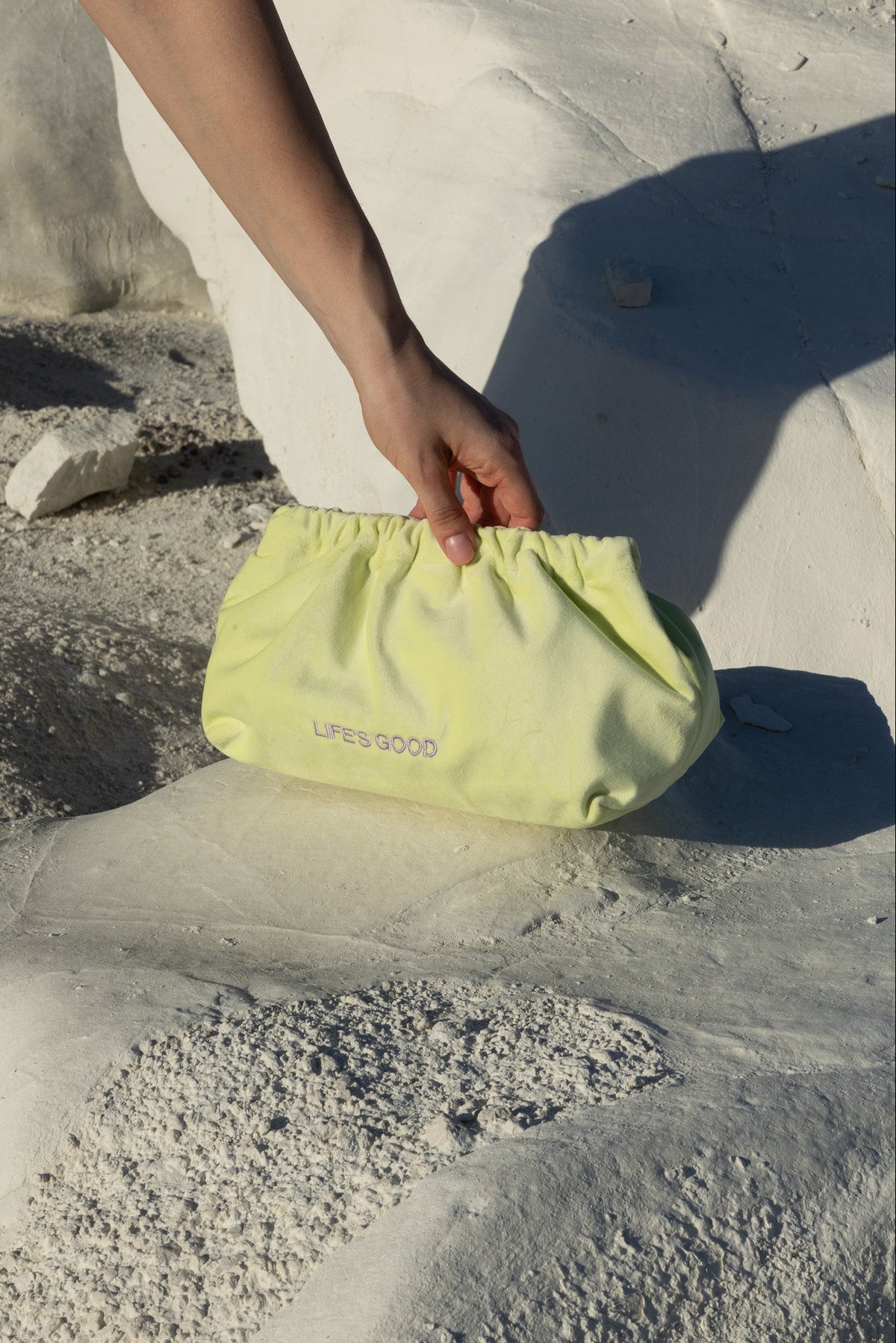 A pale yellow drawstring pouch with 'LIFE'S GOOD' printed in pink on the front, held by a person's hand against a bright white stone background.