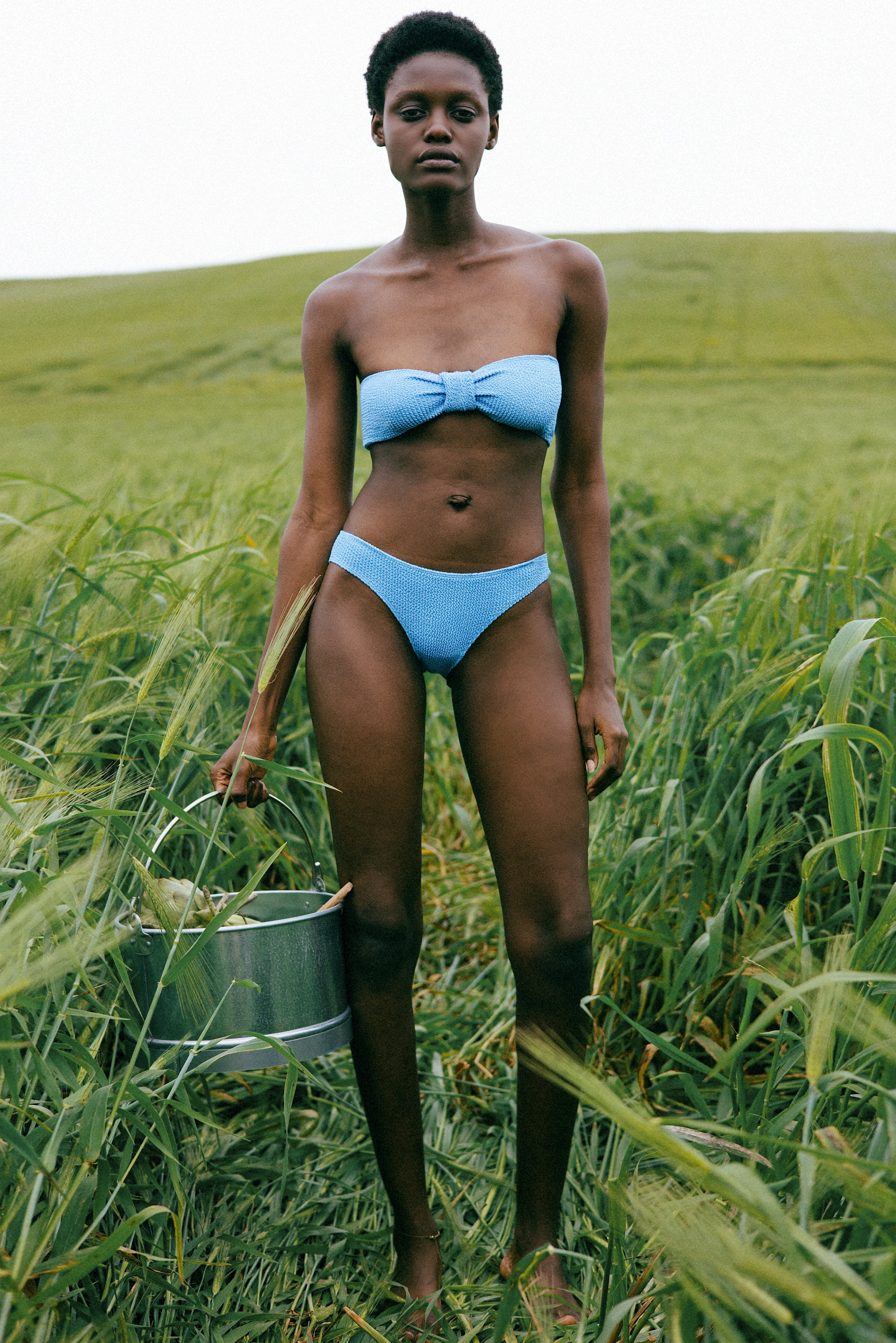 A model stands in a field of tall green grass wearing a light blue strapless bikini and holding a metal bucket.