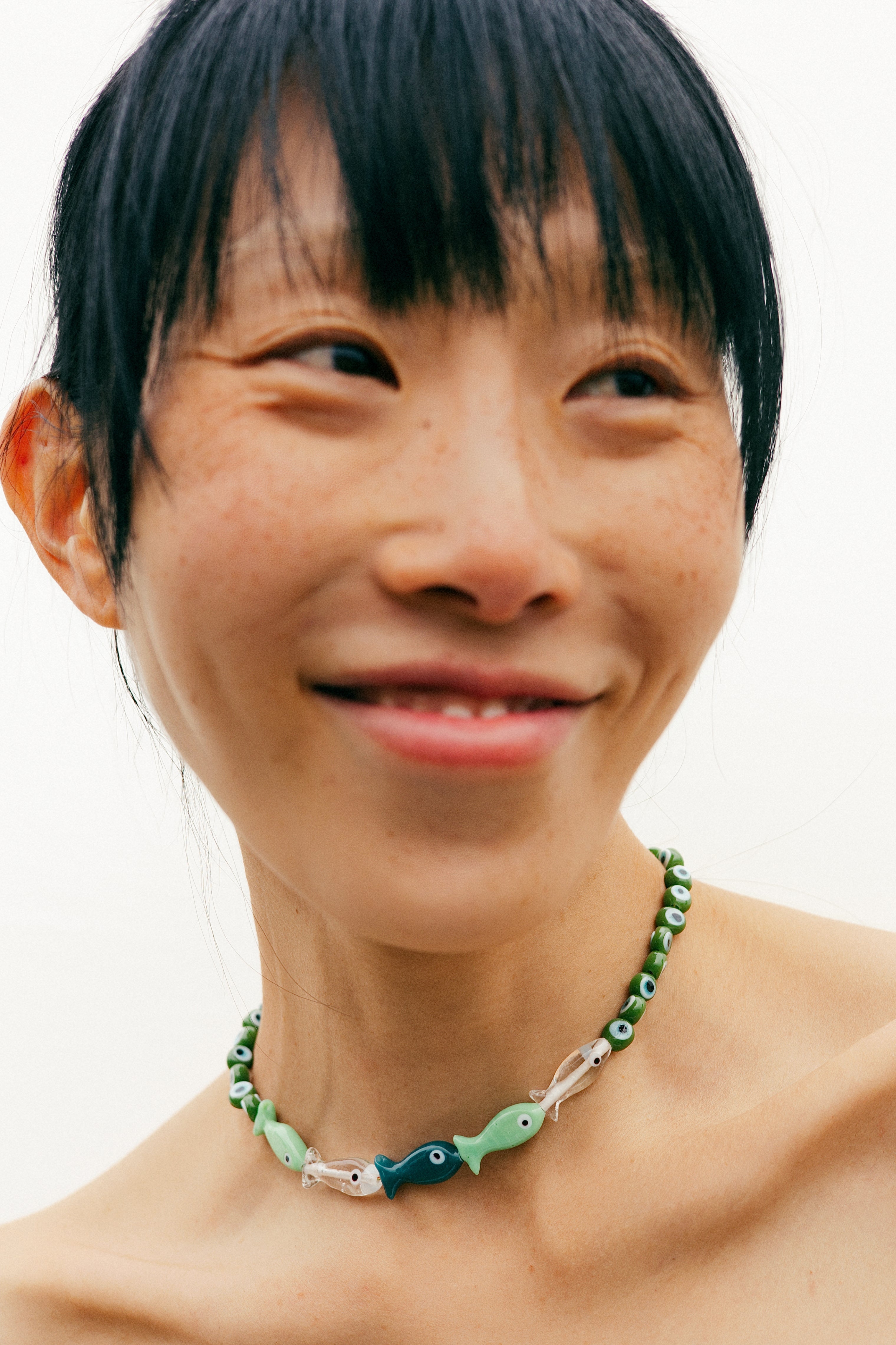 A close-up lifestyle shot of a smiling woman with short black hair wearing a green beaded necklace with fish charms against a plain white background.
