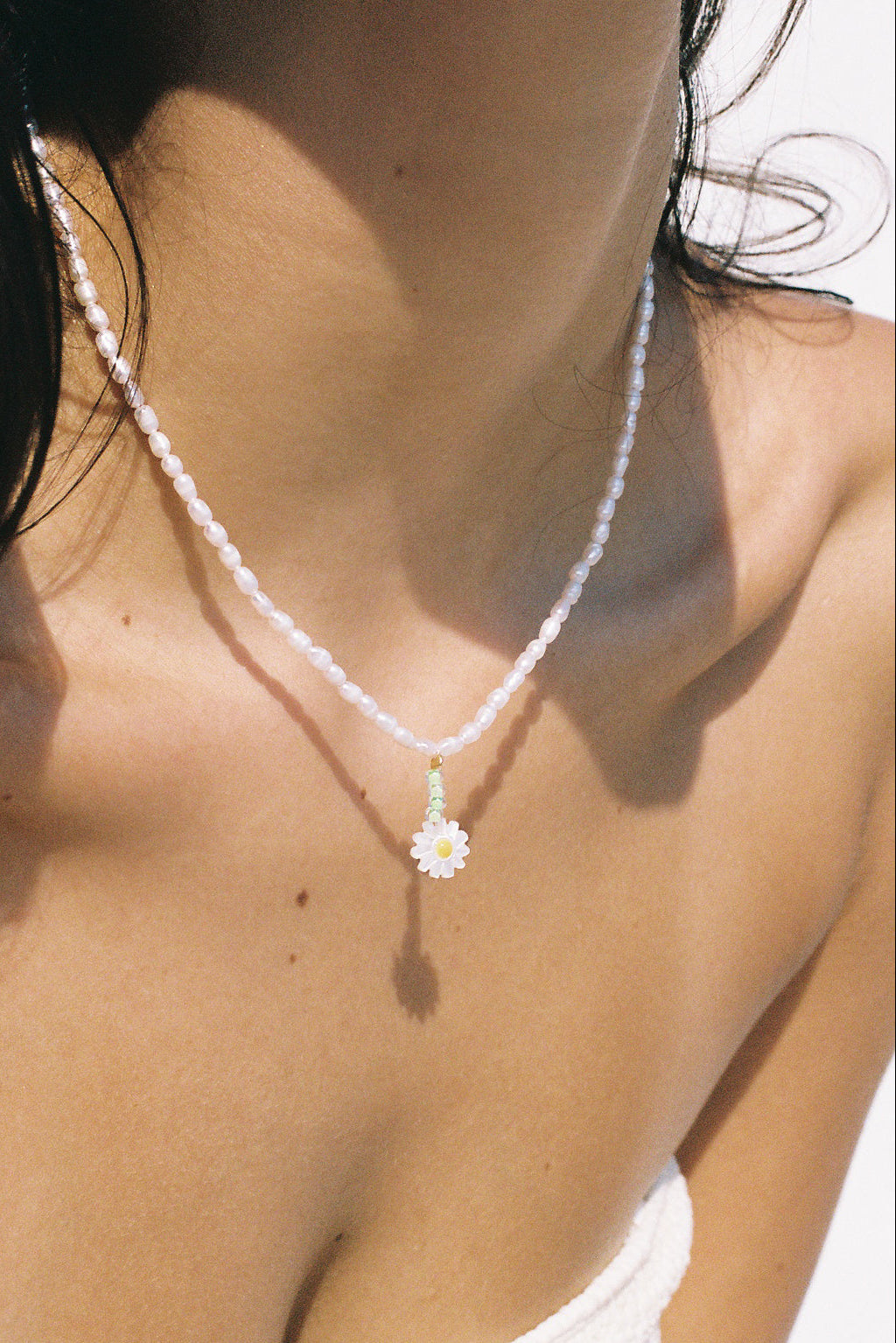 A close-up lifestyle shot of a model wearing a delicate white beaded necklace with a small white daisy pendant with a green stem.