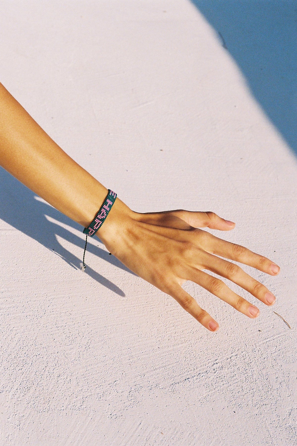 A close-up of a person's hand and wrist wearing a black beaded bracelet with colorful lettering, resting on a textured white surface under bright sunlight.