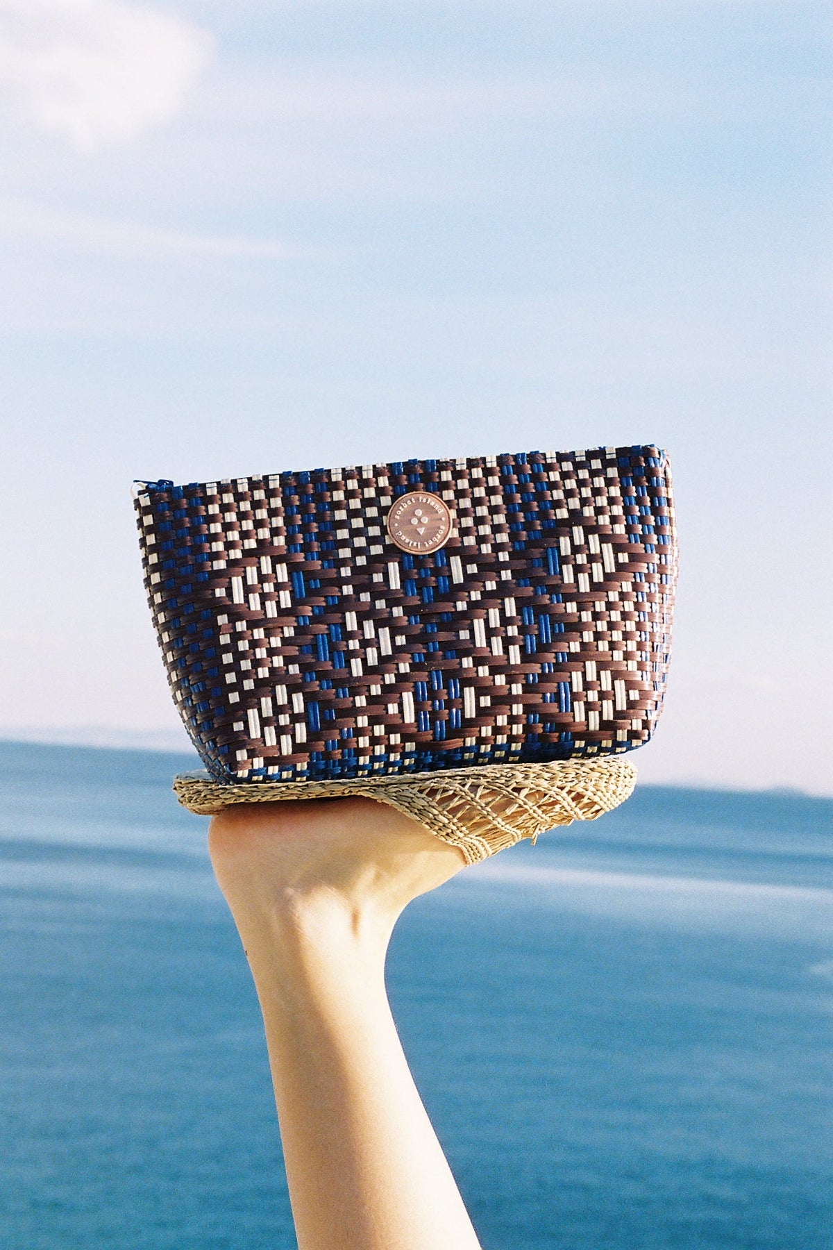 A woven clutch bag with a geometric pattern in brown, blue, and white, held up by a person's hand against a scenic background of the blue sea and sky.