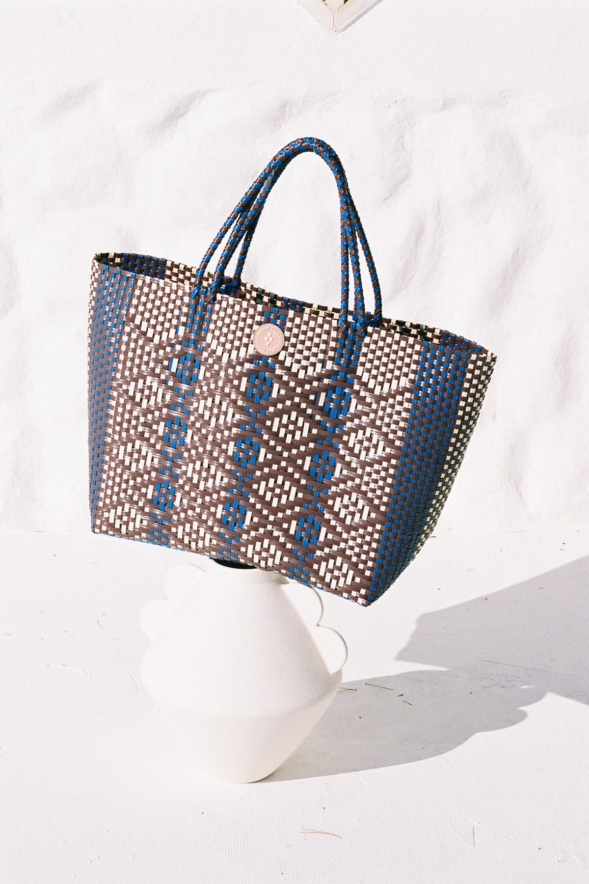 A woven tote bag with a brown and blue geometric pattern, displayed on a white ceramic stand against a textured white wall.