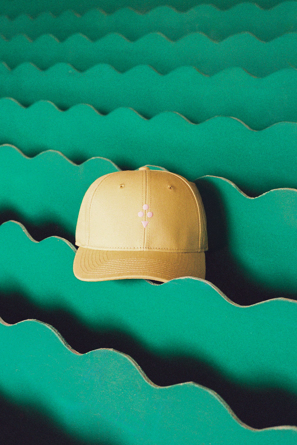 A beige baseball cap with a small pink embroidered cross logo on the front, resting on a green wavy cardboard background.