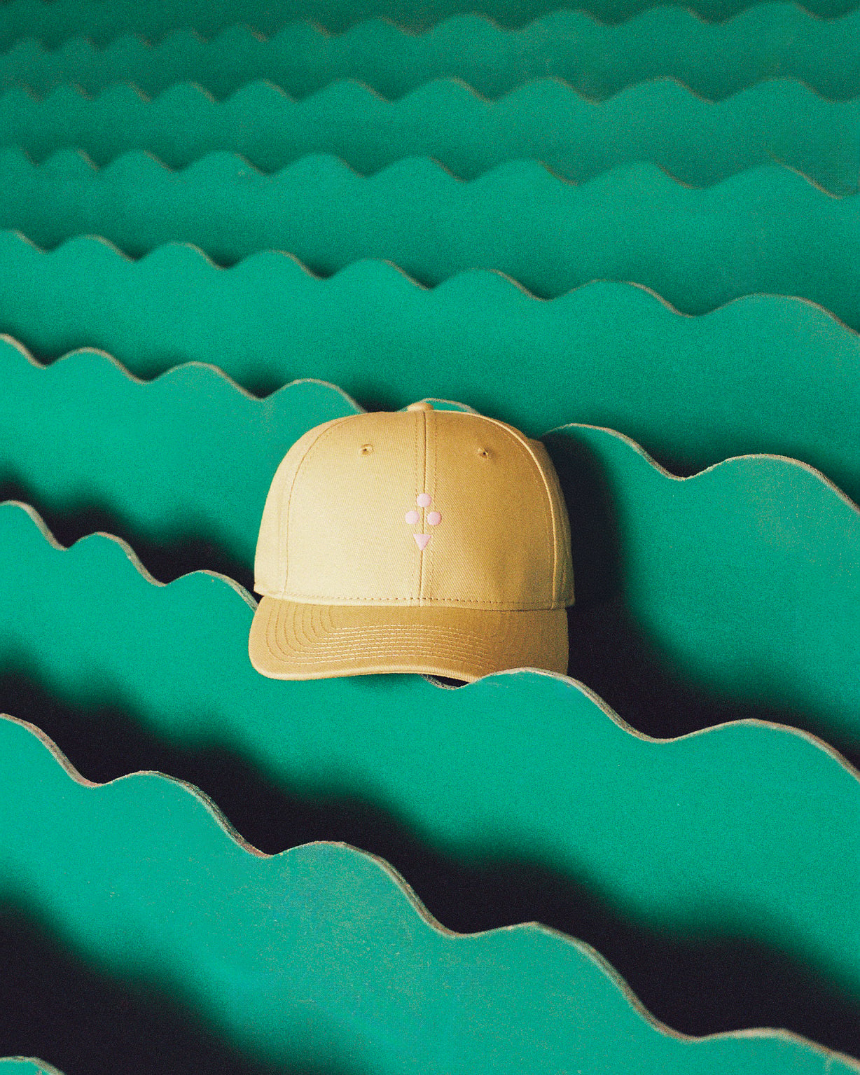 A beige baseball cap with a small pink embroidered cross logo on the front, resting on a green wavy cardboard background.