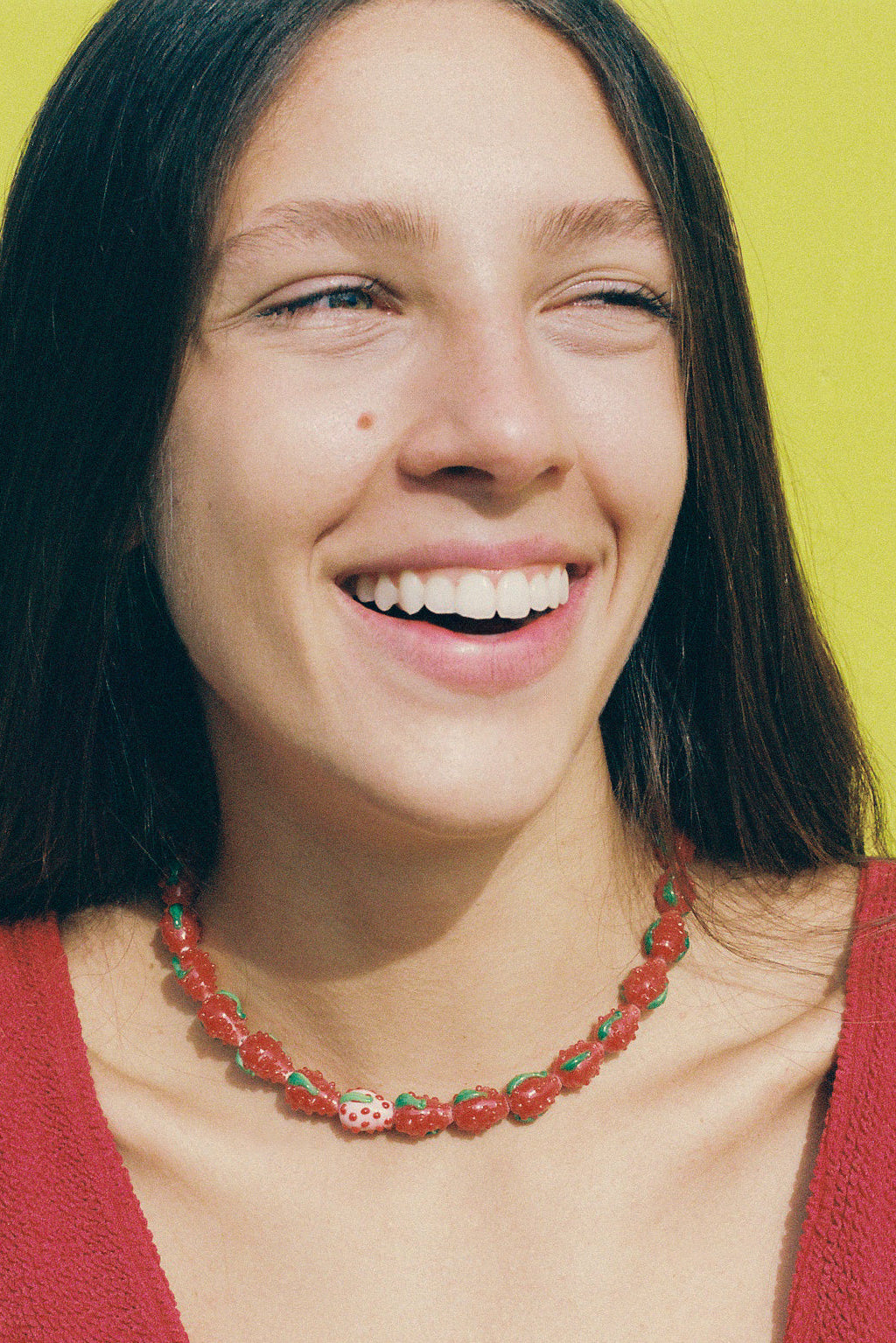 A smiling woman with dark hair wearing a red knit top and a strawberry-themed beaded necklace, set against a bright yellow background.