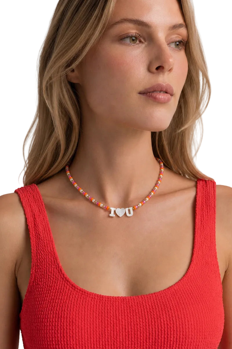 A close-up of a woman wearing a colorful beaded necklace with a silver 'I <heart> U' pendant, paired with a red ribbed tank top against a light gray background.
