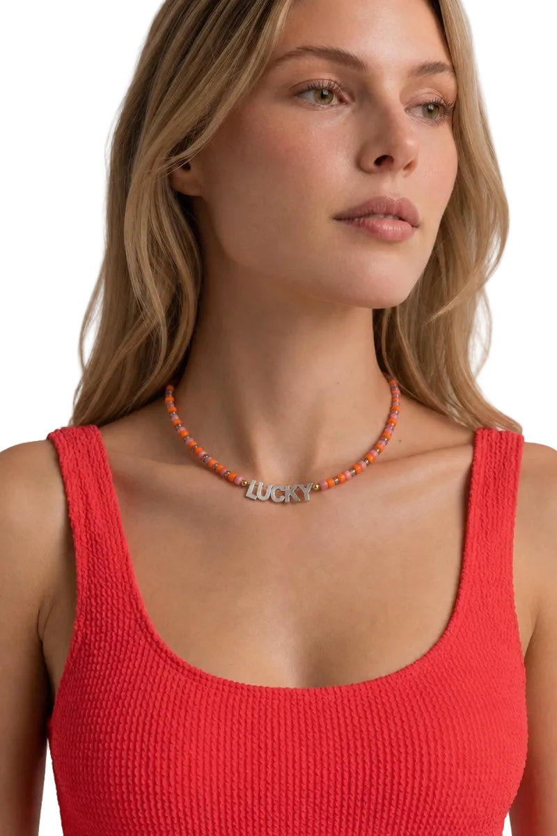 A close-up of a woman wearing a beaded orange and white necklace with a silver 'Lucky' charm, paired with a red ribbed tank top against a light gray background.