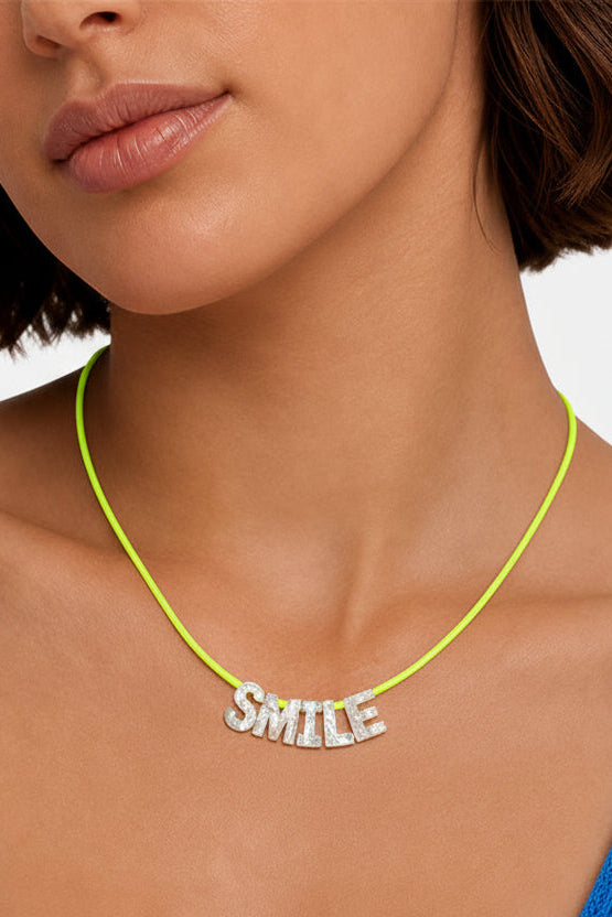 A close-up of a model wearing a bright lime green cord necklace with a silver-tone 'SMILE' word charm, paired with a blue ribbed top.