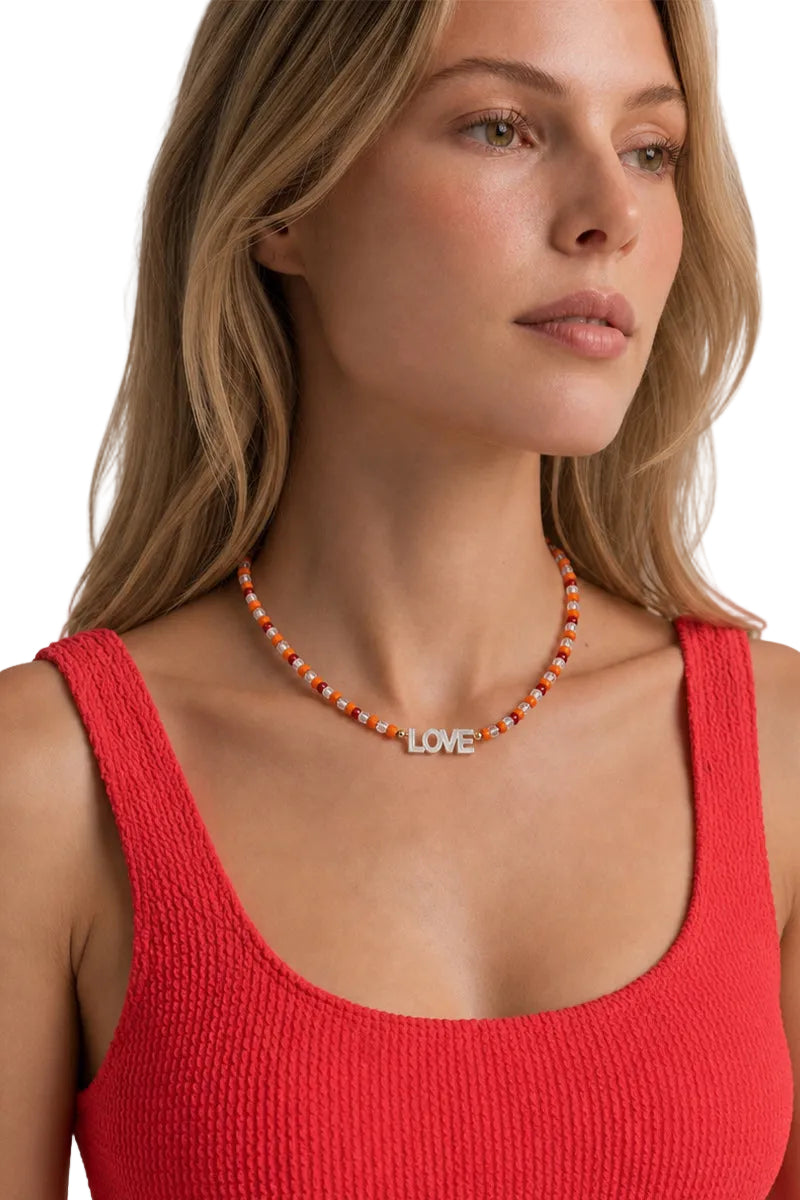 A close-up of a woman wearing a beaded necklace with a silver 'LOVE' pendant, paired with a red ribbed tank top against a light gray background.