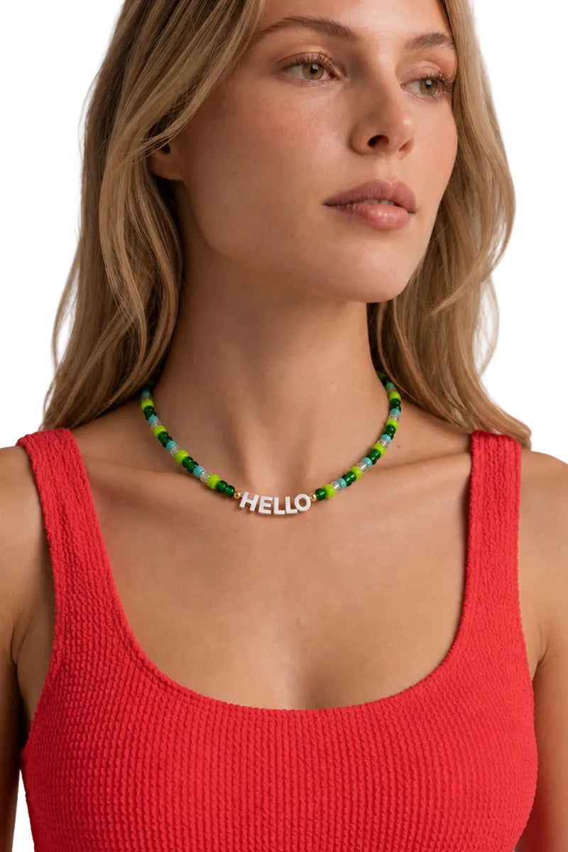 A close-up of a woman wearing a beaded necklace with green and blue beads and a silver 'HELLO' nameplate, paired with a red ribbed tank top against a light gray background.