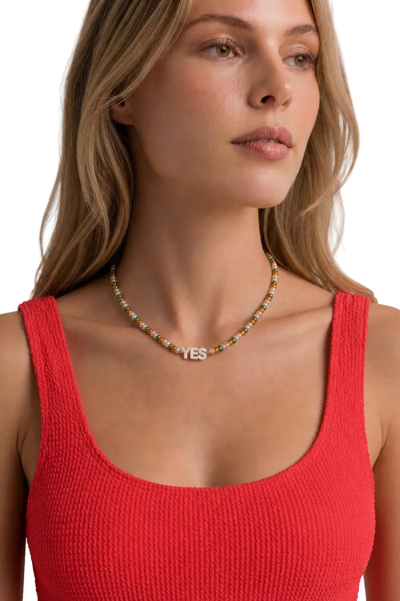 A close-up of a woman wearing a beaded necklace with a silver 'YES' pendant, paired with a red ribbed tank top against a clean white background.