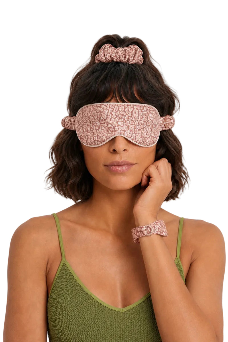 A woman wearing a pink and white textured sleep mask, matching scrunchie, and bracelet, styled with an olive green knit top against a plain white background.