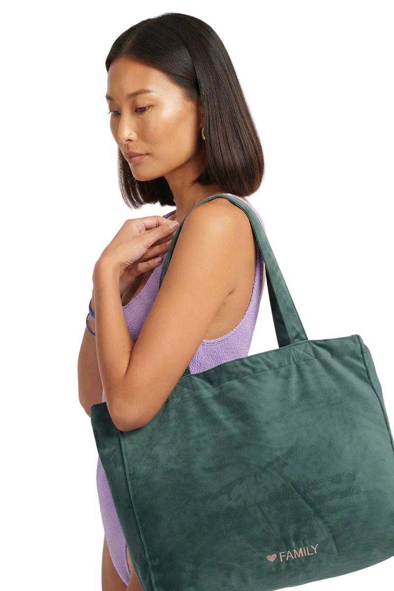 A woman wearing a purple swimsuit carries a large dark green velvet tote bag with 'FAMILY' printed on the front, set against a plain light gray background.