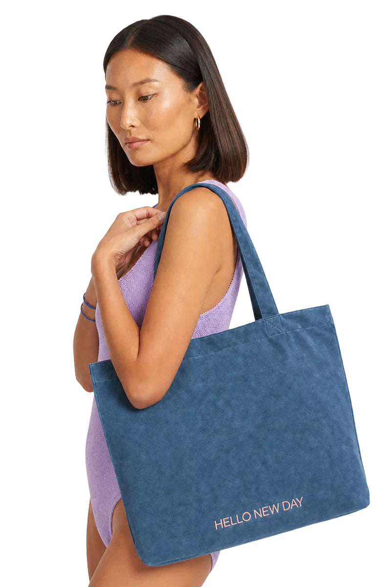 A woman wearing a lavender swimsuit carries a large blue suede tote bag with 'HELLO NEW DAY' printed in white on the bottom right corner, set against a plain light gray background.